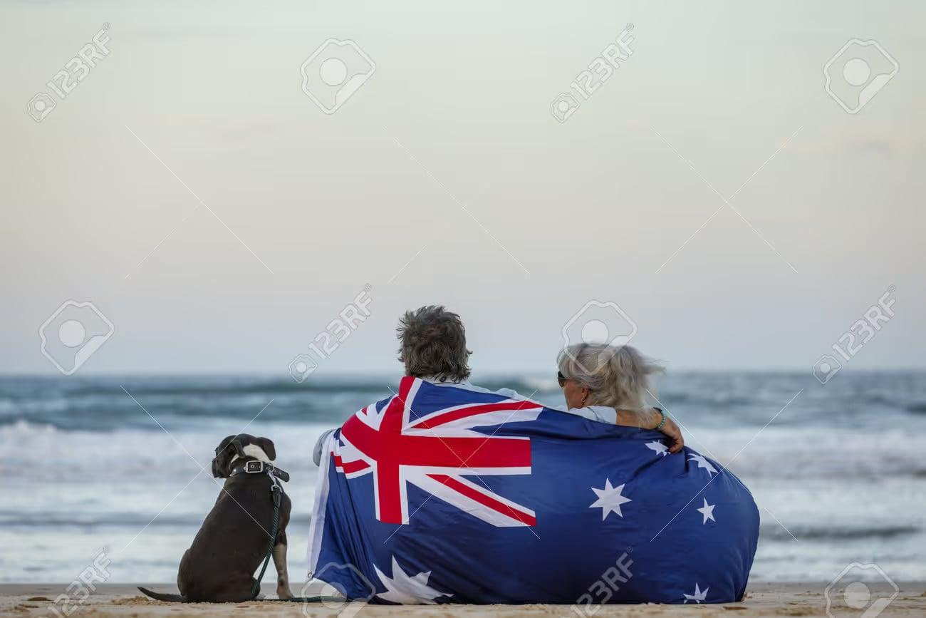 A couple wrapped in the Australian flag sit on the beach with their dog, looking out to sea